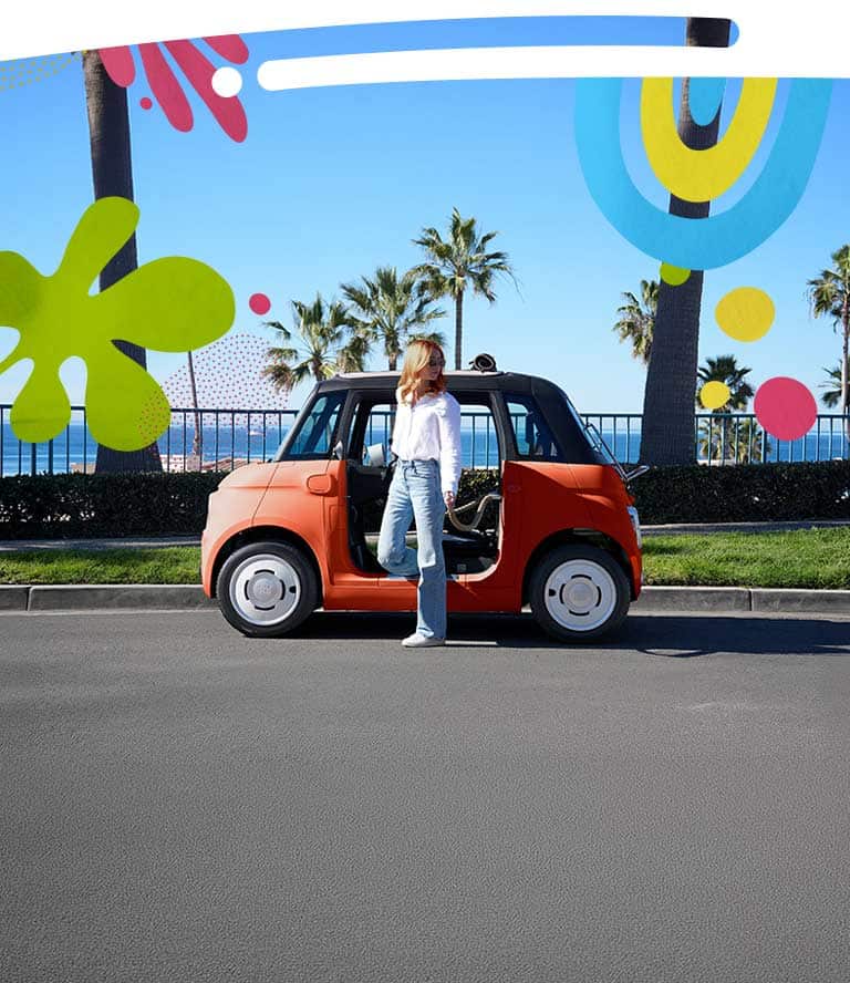A woman stands in the doorway of an Corallo Orange 2027 Fiat Topolino Dolce Vita parked along a beachside road with palm trees in the background.