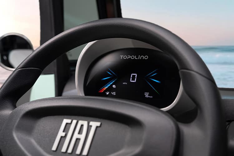 A close-up shot of the full-color Digital Cluster Display behind the steering wheel of a 2027 Fiat Topolino with the ocean visible through the windshield behind it.
