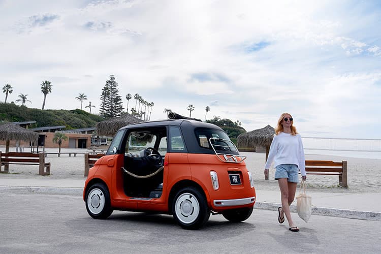 A woman walks away from a Corallo Orange 2027 Fiat Topolino Dolce Vita parked in a lot along a beach community, the day ahead of her full of possibility.