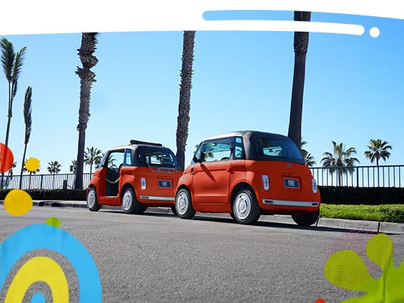 A Corallo Orange 2027 Fiat Topolino and a Corallo Orange 2027 Fiat Topolino Dolce Vita sit parked facing one another on a road with palm trees in the background.
